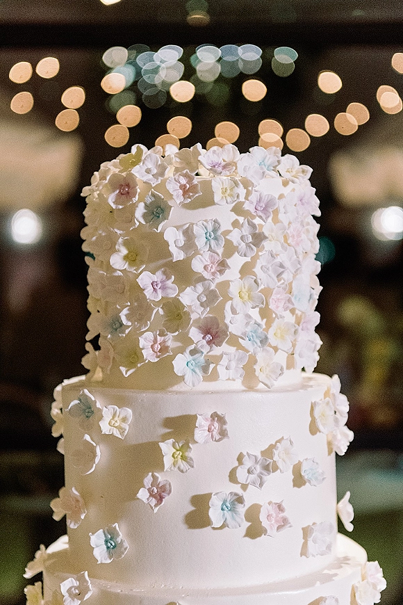 Wedding cake with white frosting and pastel sugar flowers on a stand, softly lit by string lights in a dark reception interior