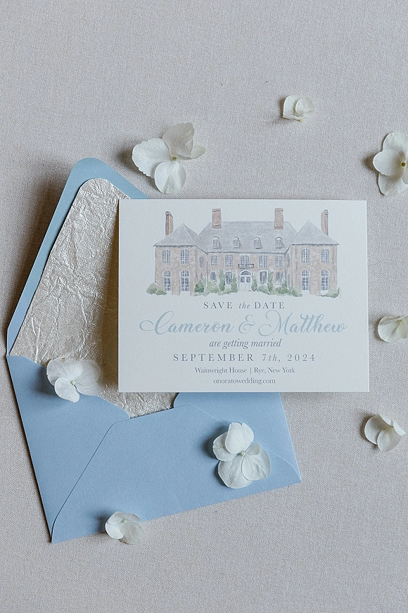 Save the date card with wedding save the date text, deckle-edge paper and watercolor venue illustration beside a blue envelope on linen fabric