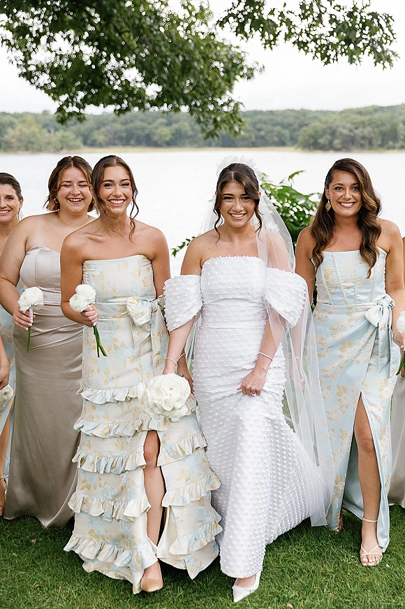 Bridal party portrait of bride with bridesmaids walking lakeside, holding white rose bouquets, in mismatched dresses with veil accents