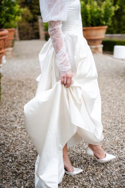 Wedding dress details with lace sleeves as bride lifts satin hem, showing engagement ring and pearl white pointed-toe heels on a garden gravel path