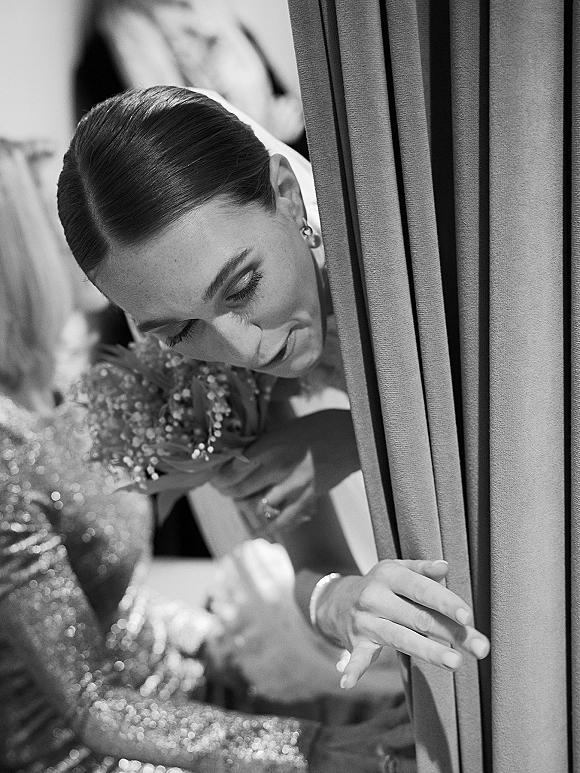 Getting ready moment as bride peeks behind curtains in window light, holding a bouquet with jewelry details and blurred figures behind