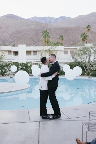 Wedding kiss portrait as groom and groom kiss in white and black tuxedos by a hotel pool, with palm trees and desert mountains beyond.