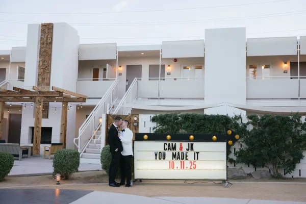 Couple portrait of two grooms standing beside a marquee sign on a courtyard staircase by the building exterior.