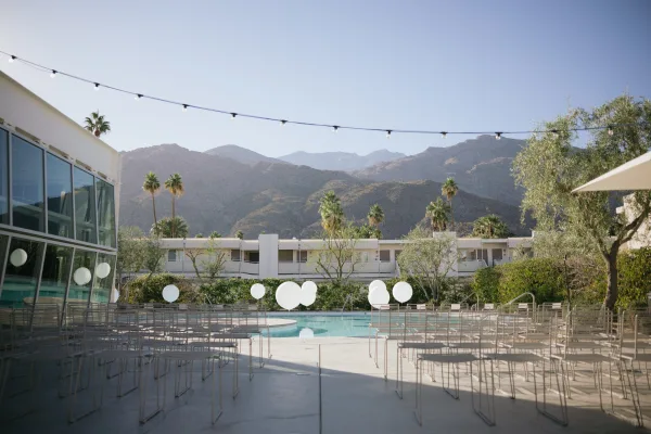 Outdoor ceremony setup for a poolside wedding ceremony with metal chairs, framed by a modern glass-walled building, palm trees, and mountains.