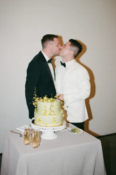 Wedding kiss portrait as two grooms kiss beside a two-tier sage green cake with yellow flowers, captured in a candid moment.