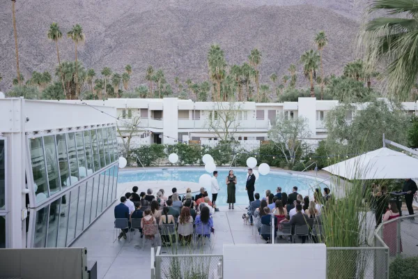 Ceremony moment at a poolside wedding ceremony, white balloons floating overhead as guests watch, with palm trees and mountains beyond.