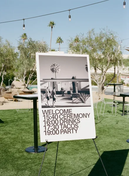 Wedding welcome sign with a black-and-white photo, set on a grass lawn under string lights and palm trees against a blue sky.