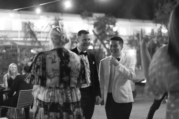 Reception moment with laughing newlyweds—two grooms—mingling on an outdoor patio under string lights against a night sky.