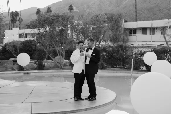 Couple portrait of two grooms in tuxedos clinking cocktails by a pool, with mountain views and a modern building framing the scene.