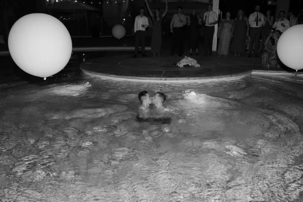 Wedding kiss moment by the night pool at a wedding pool jump reception pool party, under string lights beside the deck and patio umbrellas.