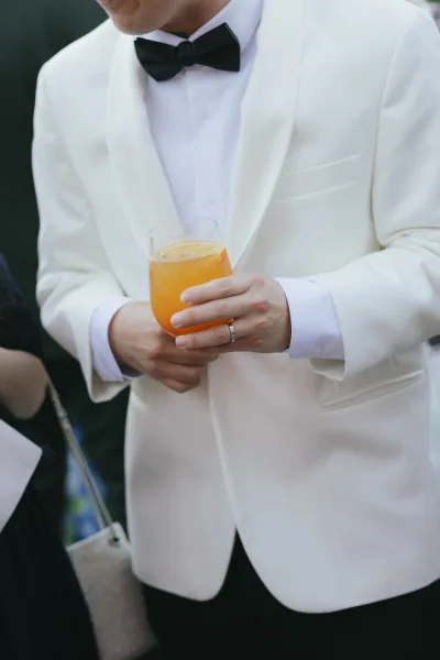 Groom attire highlighted by a white dinner jacket as he stands among guests in dark clothing, the look crisp and formal.