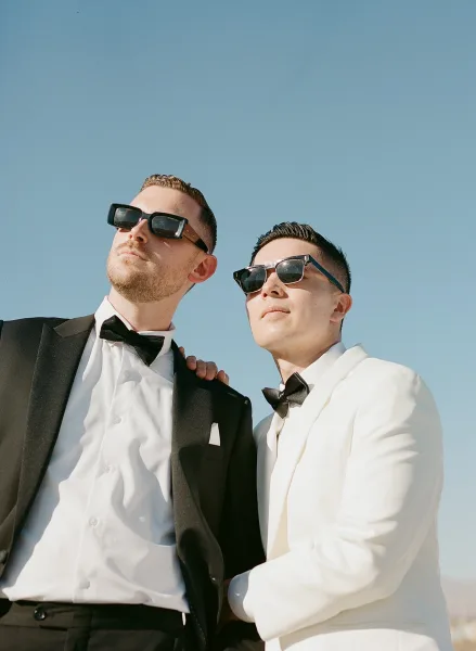 Grooms portrait of two grooms together beneath a vivid blue sky, one in a white tuxedo and one in black, both wearing sunglasses.