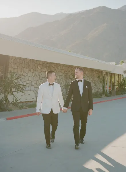 Couple portrait of two grooms, one in a white dinner jacket, against a midcentury modern building with desert plants and mountain backdrop.