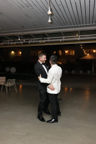 Wedding first dance as two grooms first dance beneath string lights on a covered patio, embracing by the pool against a pendant-lit facade.