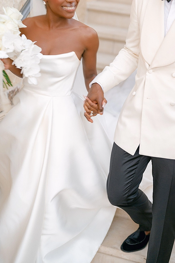 Couple portrait of bride and groom holding hands on stone steps, her veil and white bouquet visible, his white dinner jacket and bow tie