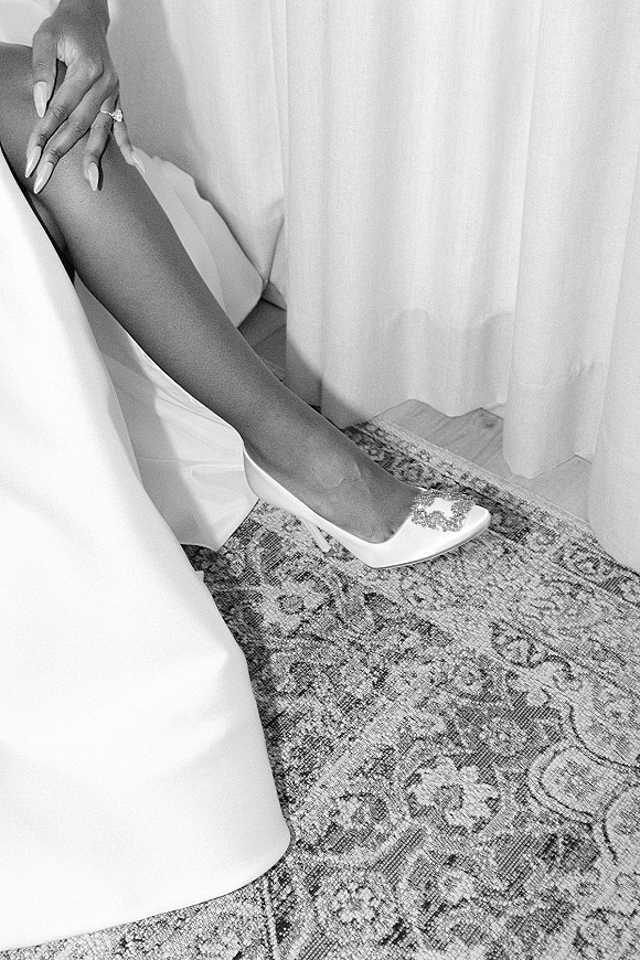Bridal shoes and wedding shoes flatlay with white rhinestone heels as a ringed hand adjusts the strap beside a bridal gown hem on a patterned rug