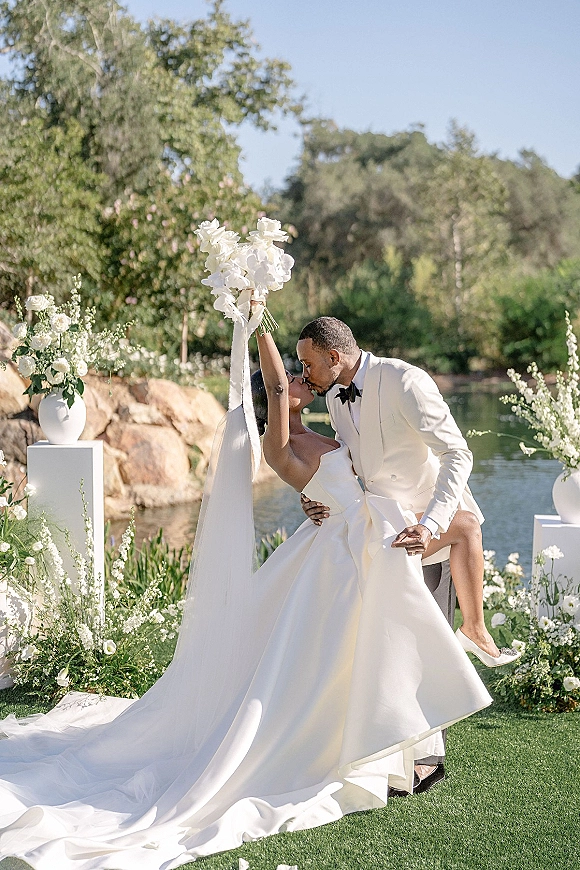 Wedding kiss portrait of groom dipping the bride as she lifts a bouquet, veil flowing, lakeside lawn with white floral pedestals behind
