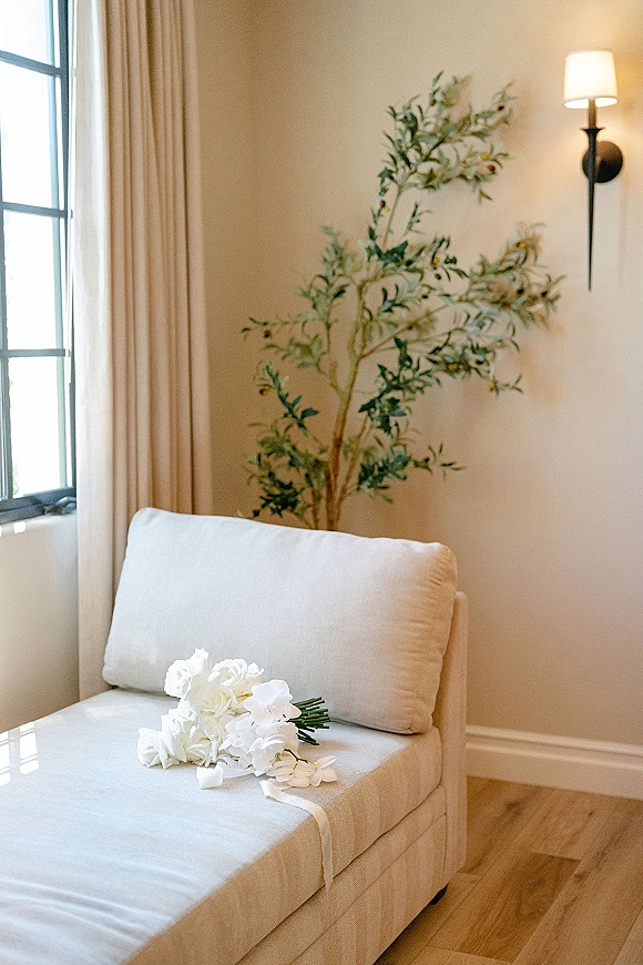 Bridal bouquet of all-white blooms with a ribbon draped over an upholstered chair by a window in a neutral bridal suite