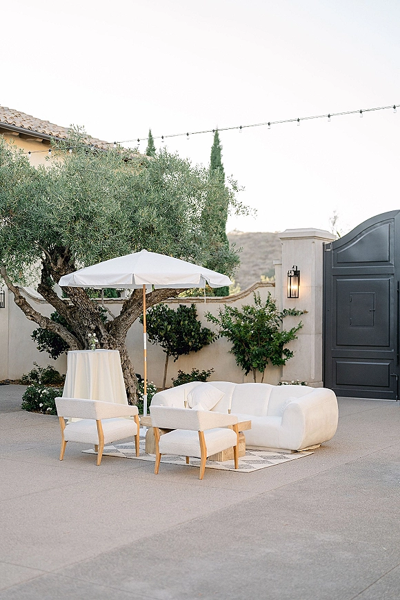 Wedding lounge seating with a white sofa, upholstered chairs, umbrella and rug, set in a stucco courtyard patio under string lights by an olive tree