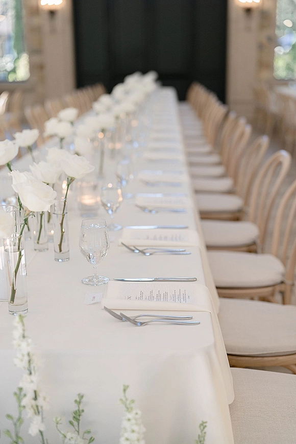 Reception tablescape on a long wedding table with white rose bud vases, glass votive candles, menus and place cards in a bright room with windows
