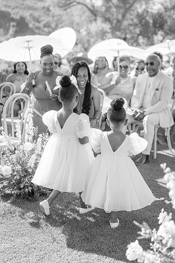 Flower girl moment as twin flower girls walking aisle in white tulle dresses, scattering petals past guests with parasols in a garden ceremony