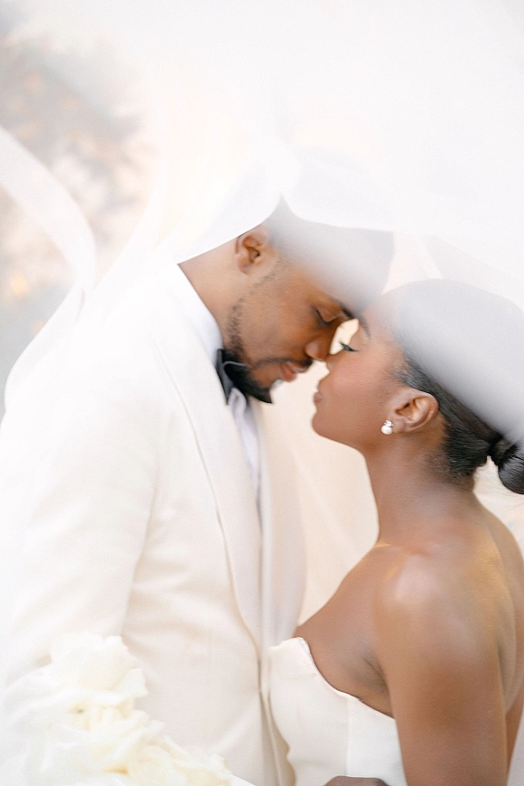 Wedding couple portrait of bride and groom in a veil, forehead touch and nose kiss, white dress and tux jacket against a bright neutral backdrop