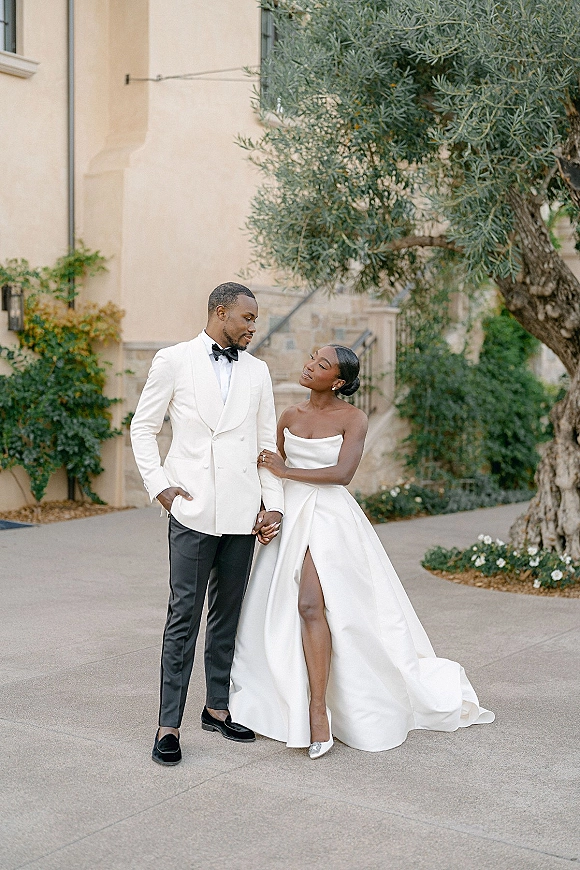Couple portrait of bride and groom holding hands on villa stone steps, strapless thigh-slit gown with long train beside an olive tree
