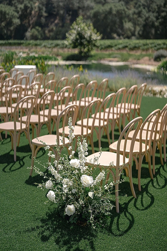 Ceremony seating with outdoor ceremony chairs lined on a lawn, featuring a white rose and greenery ground arrangement beside the aisle near a pond
