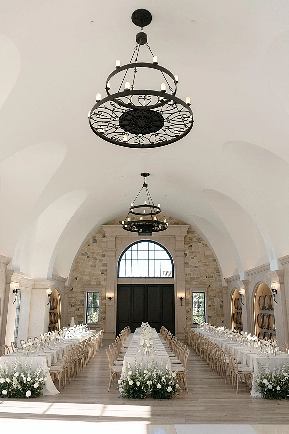 Reception tablescape with a long banquet table setup, white linens, greenery garlands, floral centerpieces, and chandeliers in a stone hall