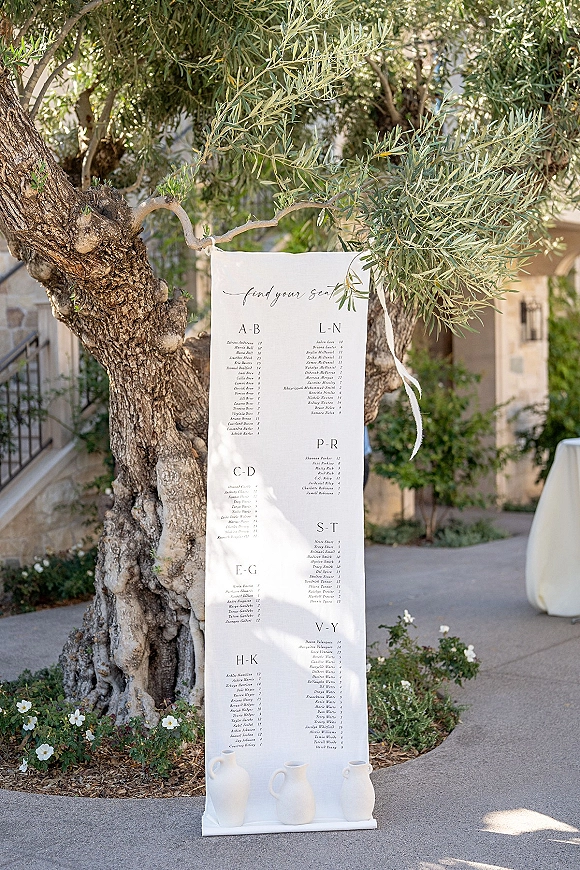 Wedding seating chart alphabetical sign in calligraphy on a white pedestal, tied with a ribbon along a garden walkway by an olive tree