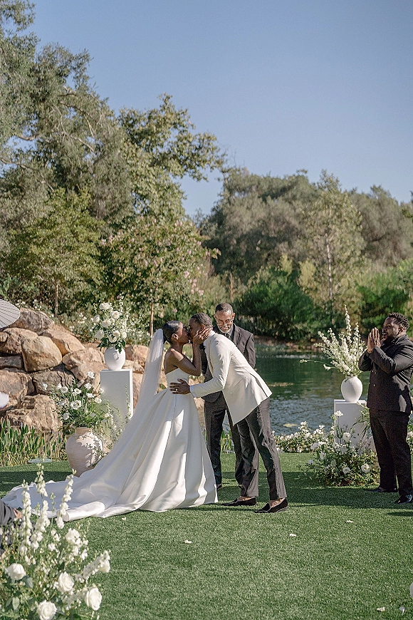 Ceremony kiss at an outdoor wedding ceremony, bride in strapless gown and veil with groom in suit before lakeside florals on pedestals