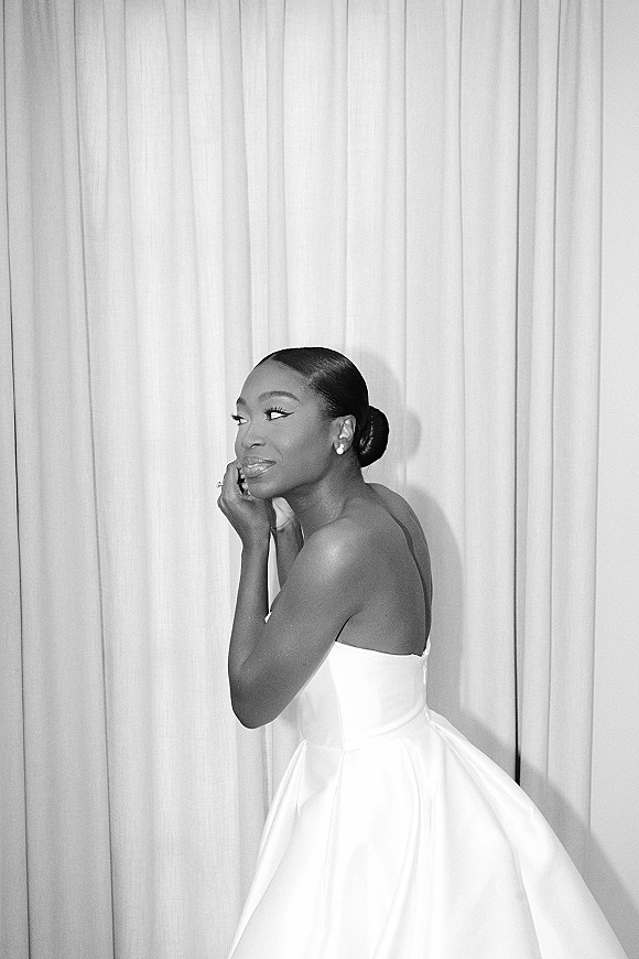 Bridal portrait in black and white of a bride in side profile, wearing a strapless dress with low bun and stud earrings by draped curtains