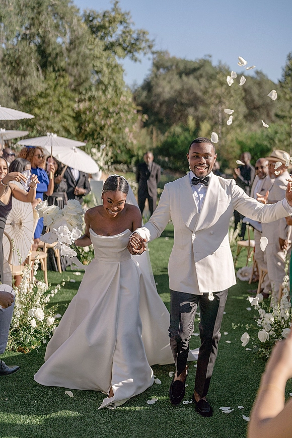 Wedding recessional as bride and groom walk the aisle holding hands, rose petals in the air, guests with white parasols in a sunny garden