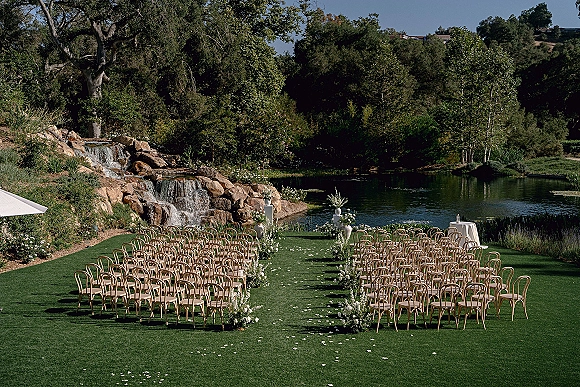 Ceremony setup with outdoor ceremony seating, bentwood chairs lining a petal-strewn aisle with white florals by a pond and waterfall