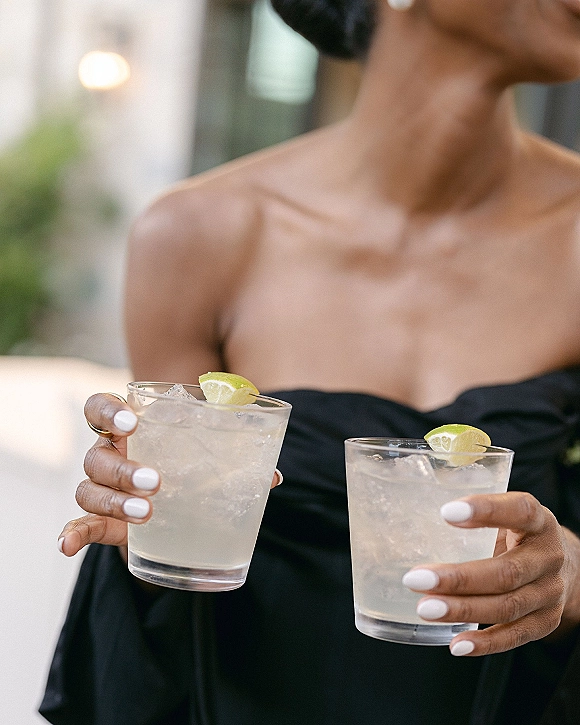 Wedding cocktails in rocks glasses with ice and lime wedge garnish, held by a bridesmaid in a black dress on a patio with blurred lights