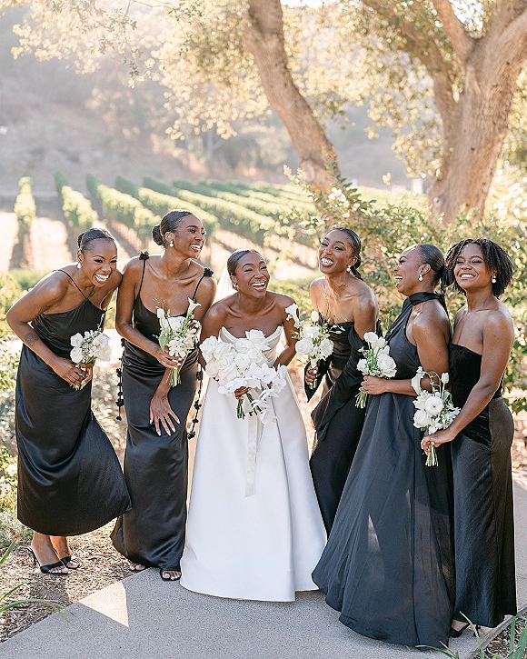Bridesmaid group portrait with bride with bridesmaids in black dresses holding white bouquets on a sunlit vineyard pathway with hills behind
