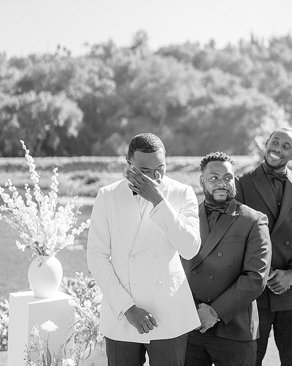 Groom portrait of a groom wiping tears in a white tuxedo jacket with boutonniere, groomsmen behind him on a hillside outdoors