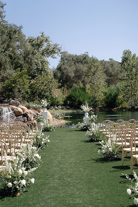 Ceremony aisle decor with white floral arrangements lining a green turf runner, wooden chairs set beside a pond and waterfall backdrop