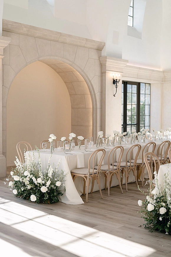 Reception tablescape with long banquet table wedding styling, white floral centerpieces and bud vases beneath a stone archway indoors