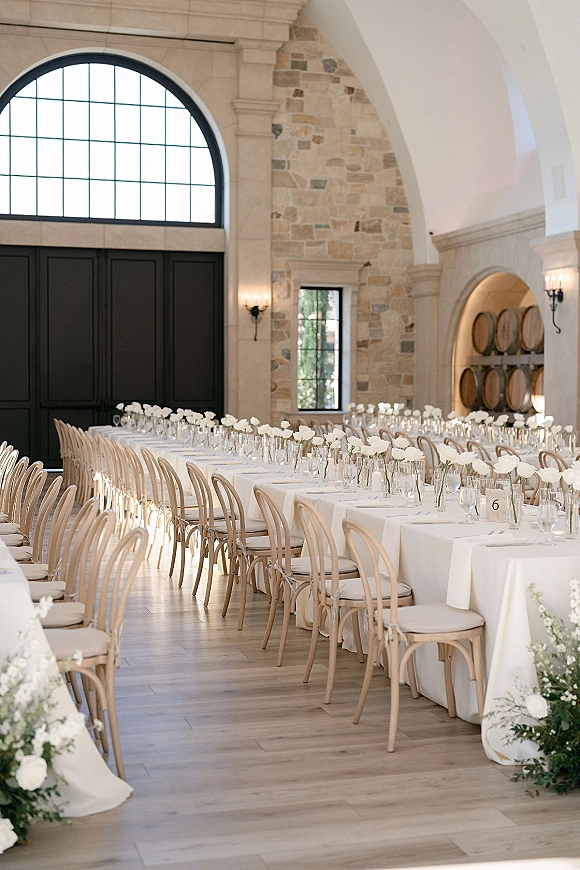 Reception tablescape with a long banquet table setup, white linens and floral centerpieces in clear vases beneath arched windows and stone walls
