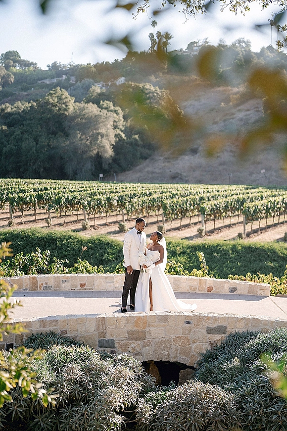 Couple portrait in a vineyard wedding portrait, bride in strapless dress with veil holding bouquet beside groom in white tux on a stone terrace