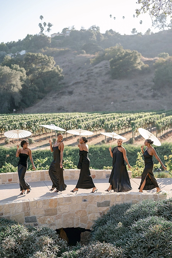 Bridesmaid portraits with parasols show women in black bridesmaid dresses holding white parasols on a sunny vineyard patio by a stone bridge