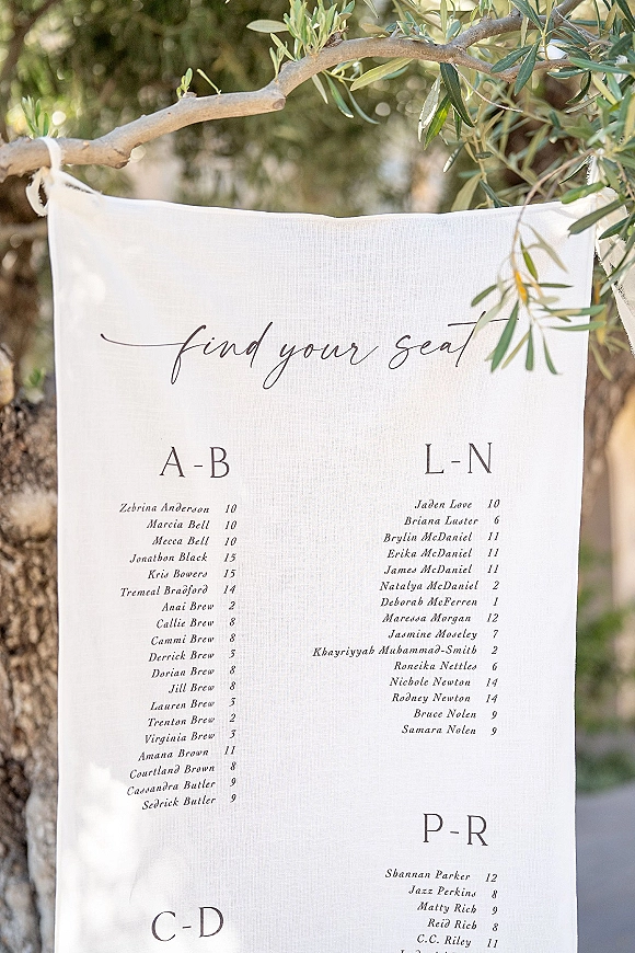 Wedding seating chart linen find your seat sign with black calligraphy text hanging from a tree branch amid lush garden foliage