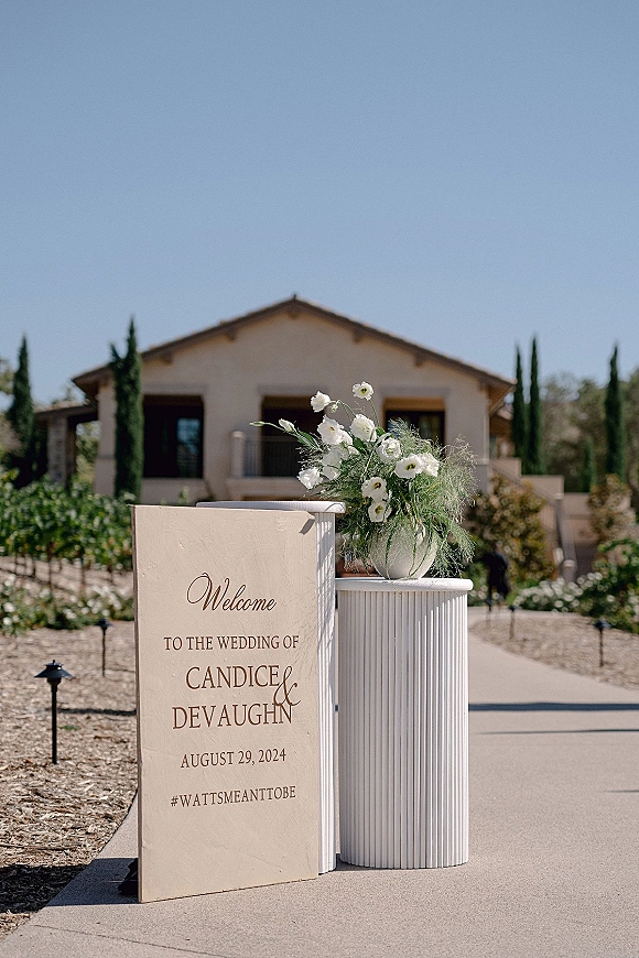 Wedding welcome sign on a pedestal with white flowers and greenery in a vineyard walkway, with trees, house facade, and blue sky behind