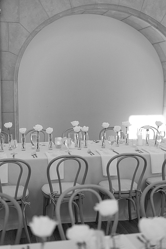 Reception tablescape on a long head table with white rose bud vases, candlelight, menus, and glassware set before an arched wall