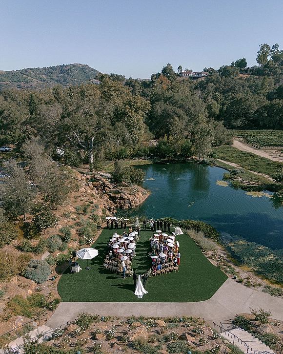 Outdoor ceremony setup with lakeside wedding ceremony chairs in a circle, white umbrellas and aisle runner on green lawn by the lake and vineyard rows