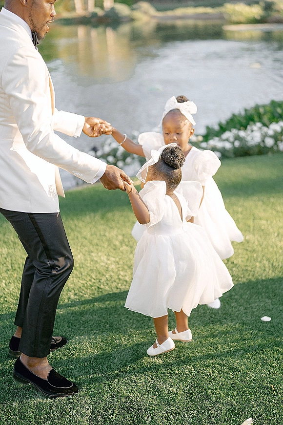 Wedding flower girls holding hands in white tulle dresses with hair bows, walking on a lawn by a lake with greenery and white flowers