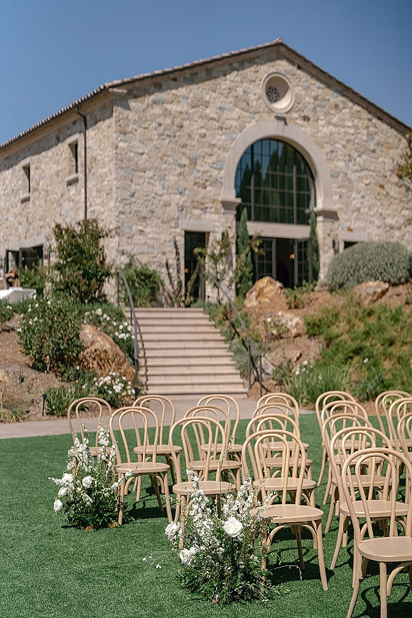 Ceremony seating with outdoor wedding ceremony chairs, curved wood chairs lining an aisle with white roses and greenery before a stone building
