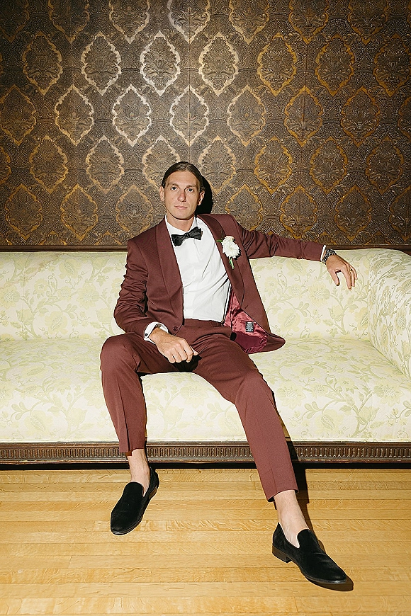 Groom portrait in a burgundy tuxedo with black bow tie and boutonniere, seated by a vintage sofa against patterned wallpaper