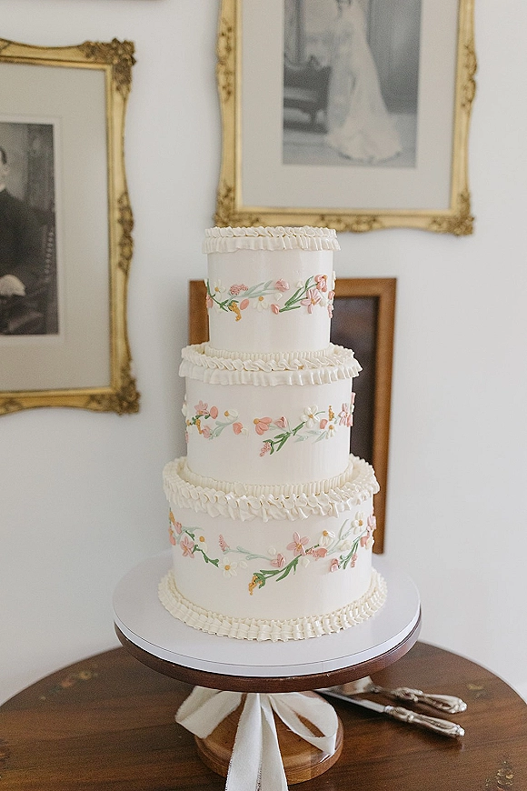 Wedding cake with hand painted floral icing on a stand with white ribbon, ruffle frosting, knife and server on wooden table by framed photos